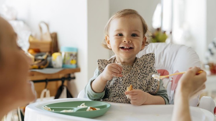 Cheerful baby girl sitting on high chair while eating meal. Female toddler is being fed by mother. They are at home.