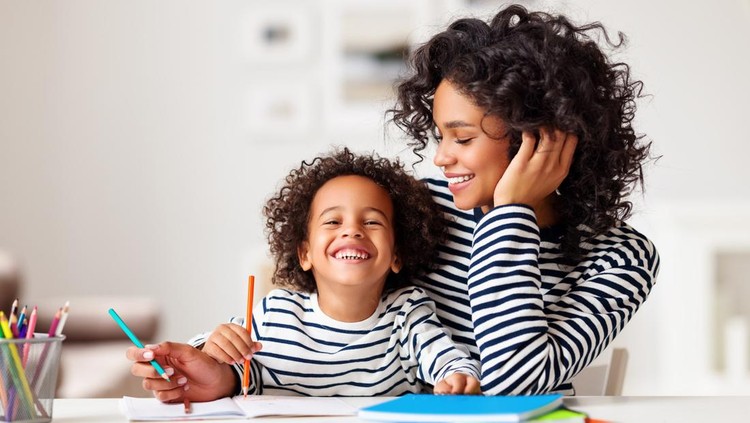 Cheerful ethnic woman laughing while helping happy boy to do homework assignment in cozy room at home