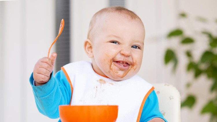 Baby boy in high chair feeding himself and making a face