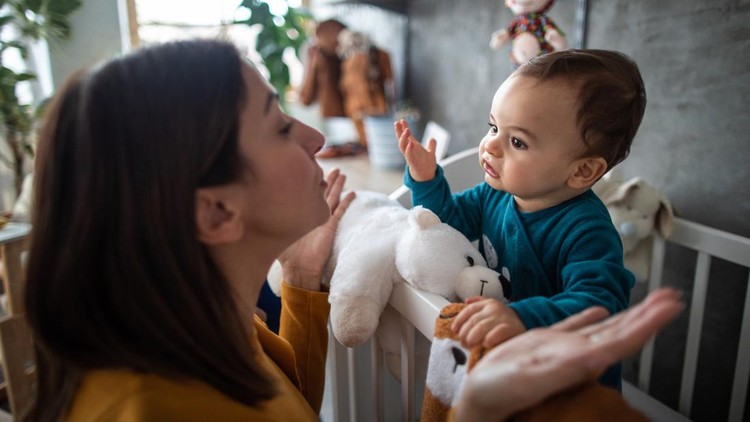 Cute baby boy in pajamas trying to explain something to his mother while standing in crib early in the morning