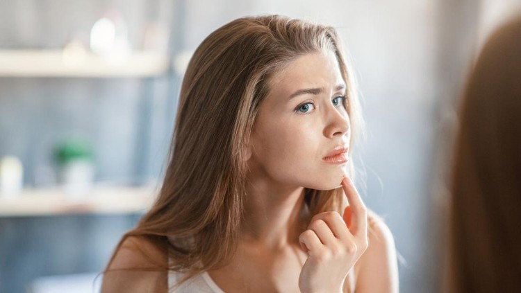 Upset millennial girl touching pimple on her chin in front of mirror at home