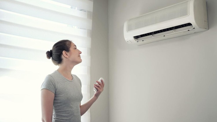 Ilustrasi Penggunaan AC Young brunette woman in her modern apartment. She turns on the air conditioner from the remote control. Climate control at home with split system.