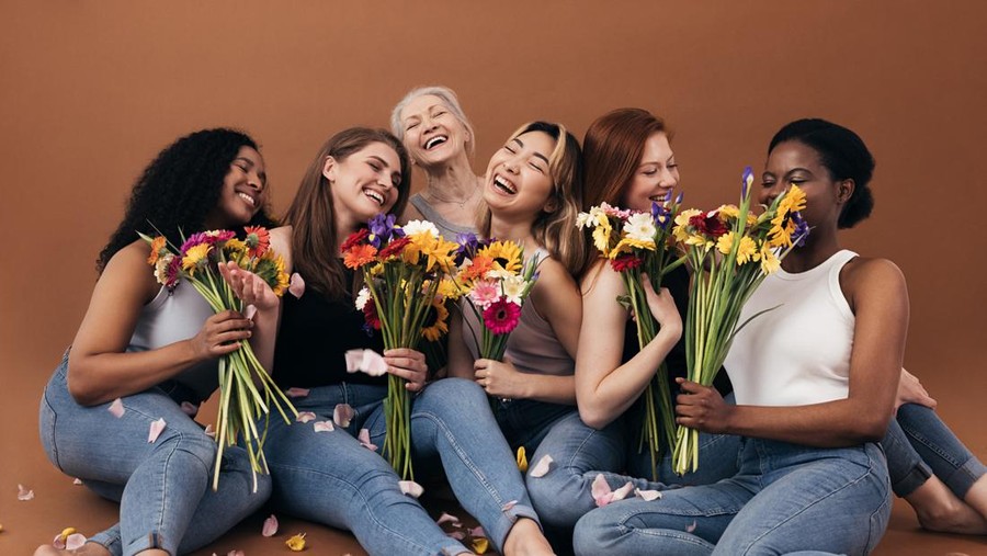 Six women of different ages and body types holding bouquets while sitting in studio against a brown background