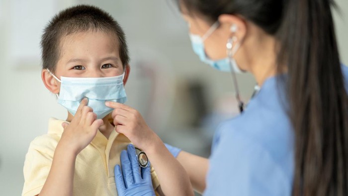 A sweet Asian boy sits up on the doctors exam table in casual clothing as he smiles behind his masked face.  His female doctor is listening to his heart with her stethoscope as she smile down at him behind her mask.  Both are wearing personal protective equipment to keep them safe from viruses.