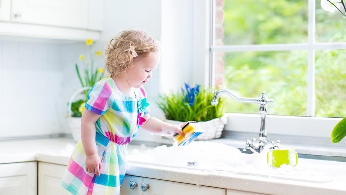 Cute curly toddler girl in a colorful dress washing dishes, cleaning with a sponge and playing with foam in the sink in a beautiful sunny white kitchen with a garden view window in a modern home