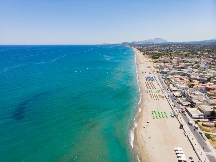 Aerial images from a drone of the coast of Rethymno town with the long beach and the beach bars in Creta island. People are seen enjoying the sun under the umbrella at the beach bar or swimming in the crystal clear sea. Rethymno is a historic Mediterranean beach town on the northern coast of Crete, laying on the Aegean Sea with a population of 40.000 people. A touristic destination with a historic Venetian port and town, Archaeological sites, endless sandy beaches, watersports, nice beautiful traditional taverns, and a big variety of hotels. Tourism is bouncing back with reservations exceeding the pre-pandemic numbers, putting Greece and the Greek islands on high demand. Tourists travel after the Covid-19 Coronavirus pandemic. Rethimno, Crete island, Greece on June 13, 2022 (Photo by Nicolas Economou/NurPhoto via Getty Images)
