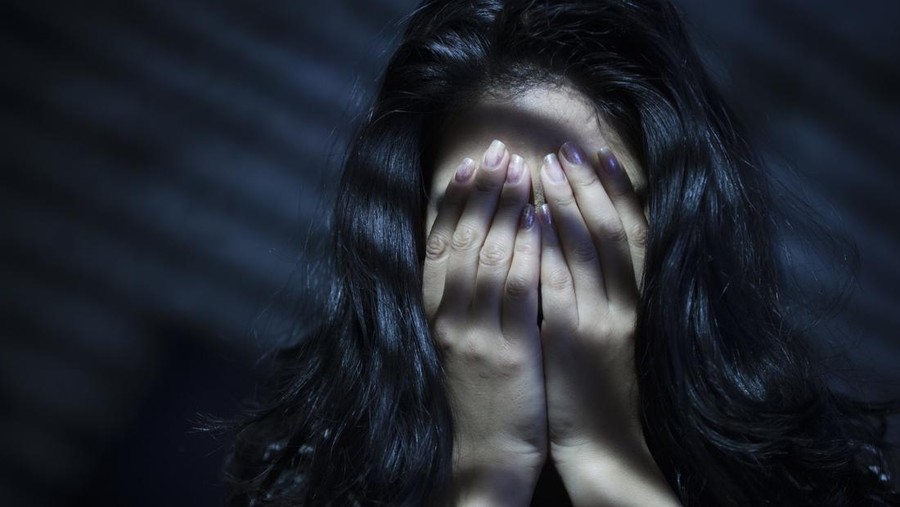 Indoor image of a young woman covering her face with her both hands out of embracement and sadness. Striped Light and shadows are falling on her through blinds. One person, low key, horizontal composition with selective focus and copy space.