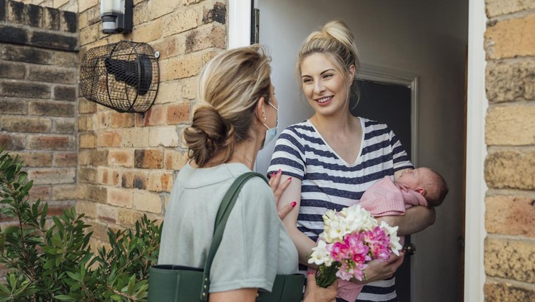 A shot of a grandmother wearing a protective face mask visiting her daughter and granddaughter with a special gift of flowers, they are greeting at the front door.