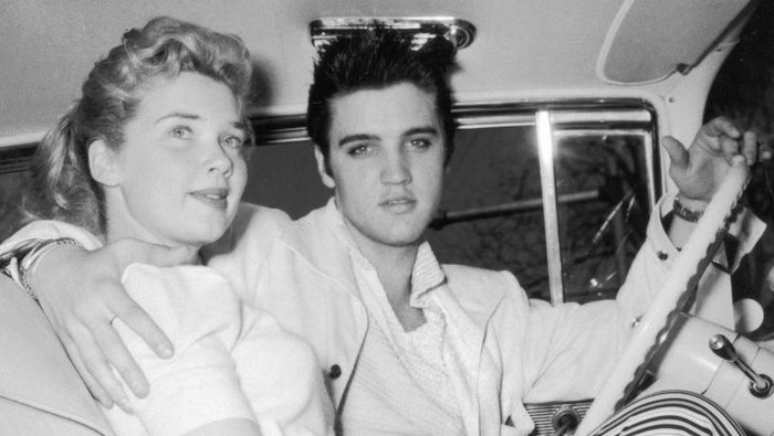 circa 1955:  American singer and actor Elvis Presley (1935-1977) sits in the driver's seat of a car with his arm around an unidentified young woman.  (Photo by Hulton Archive/Getty Images)