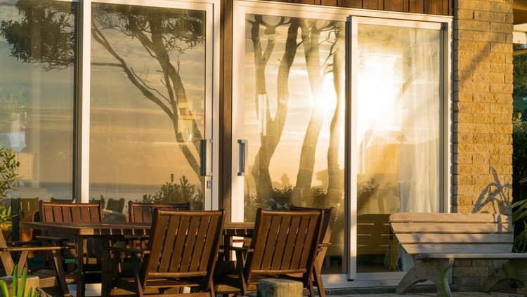 Waterfront home exterior with outdoor furniture and large windows reflecting sunrise over the sea