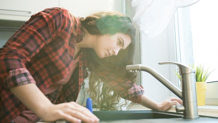 Serious concentrated young woman in checkered shirt checking faucet while having problem with dropping faucet in kitchen