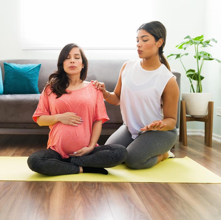 Kelas prental Sporty doula doing breathing exercises with a caucasian pregnant woman while sitting on a yoga mat in the living room