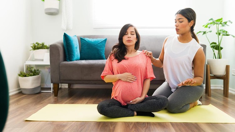Kelas prental Sporty doula doing breathing exercises with a caucasian pregnant woman while sitting on a yoga mat in the living room
