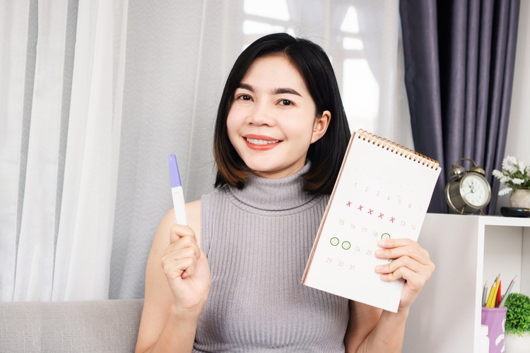 smiling Asian woman hand holding pregnancy test and calendar showing mark on date menstrual cycle planning for ovulation day