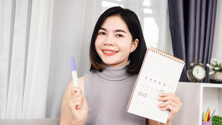 smiling Asian woman hand holding pregnancy test and calendar showing mark on date menstrual cycle planning for ovulation day