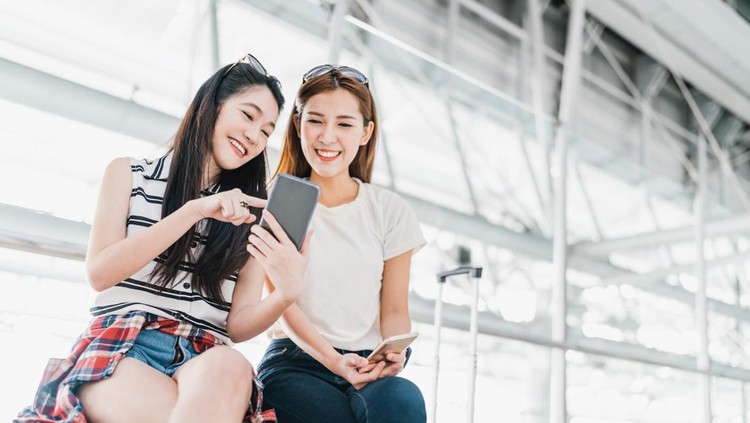 Two happy Asian girls using smartphone checking flight or online check-in at airport together, with luggage. Air travel, summer holiday, or mobile phone application technology concept