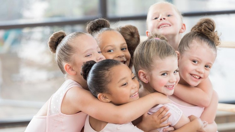A group of young smiling ballerinas hug each other in a large huddle.  Three of them are looking at the camera.