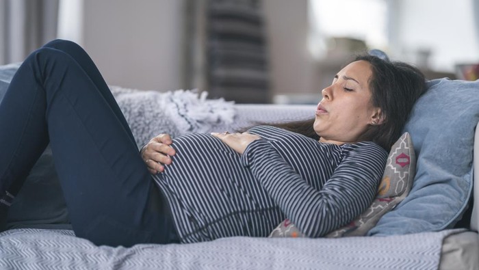 A young woman of Asian descent is pregnant. She is in her second trimester. The woman is lying on the couch in discomfort. She is breathing heavily and is resting her hand on her stomach. Prenatal, morning sickness, back pain, heartburn, and constipation concepts.