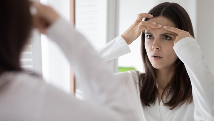Head shot mirror reflection unhappy annoyed young woman squeeze pimple on forehead, dissatisfied girl wearing white bathrobe checking face, standing in bathroom, skin problem and acne