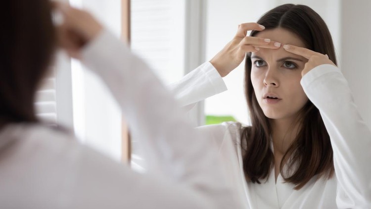 Ilustrasi Jerawat Head shot mirror reflection unhappy annoyed young woman squeeze pimple on forehead, dissatisfied girl wearing white bathrobe checking face, standing in bathroom, skin problem and acne