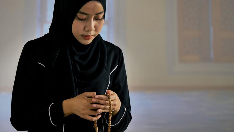 Asian Muslim young woman in black traditional hijab praying with Islamic beads in mosque.