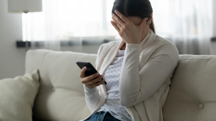 Woman sit on sofa holding smartphone cover face with hand feels scared humiliated suffering from cyberbullying being on-line abused by stalker. Bad news, life troubles, break up with boyfriend concept