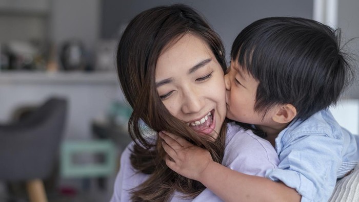 Close up shot of an adorable preschool age boy of Asian descent embracing his mom and giving her a kiss on the cheek. The loving mom is smiling with appreciation and affection. The family is spending quality time together at home.