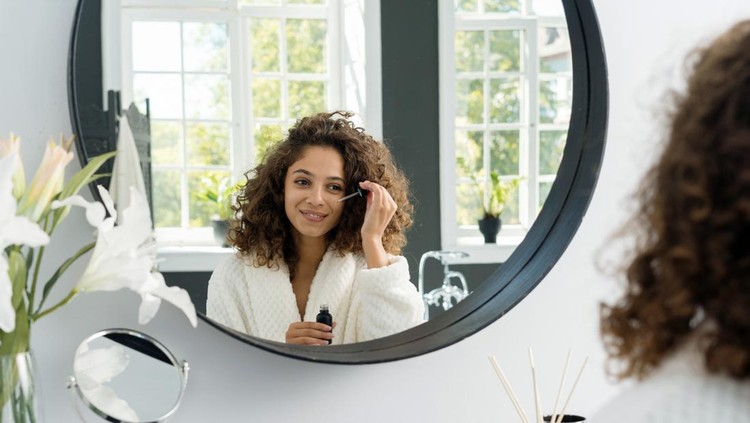 Concept of beauty procedure at home. Happy young adult african american woman holding pipette with anti wrinkle serum near eye, looking at mirror wearing in bathrobe, sitting at bathroom