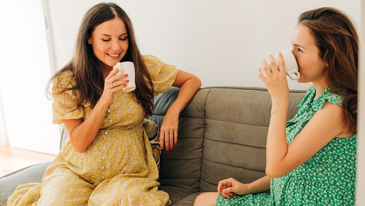 Pregnant women wearing summer dresses sitting on sofa while having a conversation over a cup of tea. Two happy women friends talking and laughing while drinking tea at home