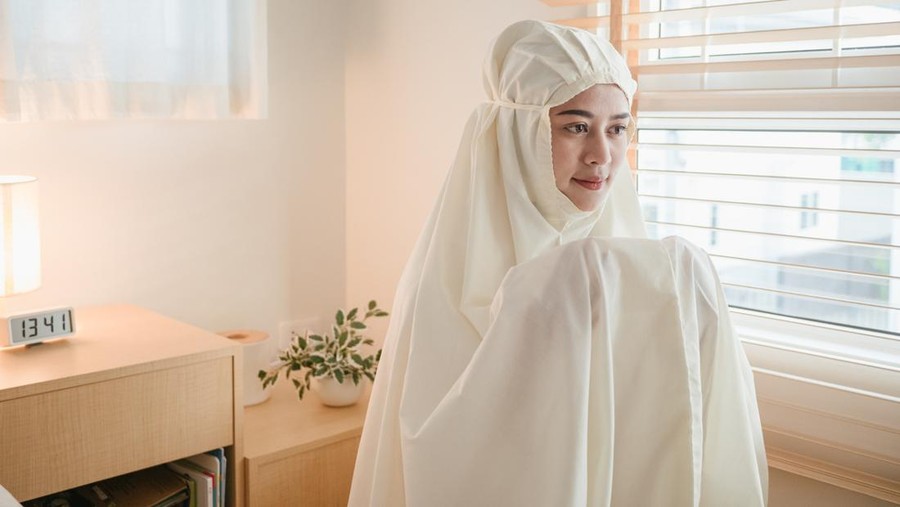 Young muslim woman in beige hijab and traditional clothes praying for Allah at home