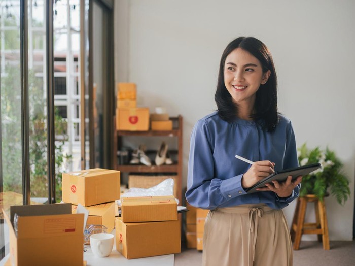 Portrait business Asian woman smile and use tablet checking information on parcel shipping box before send to customer. Entrepreneur small business working at home. SME business online marketing.