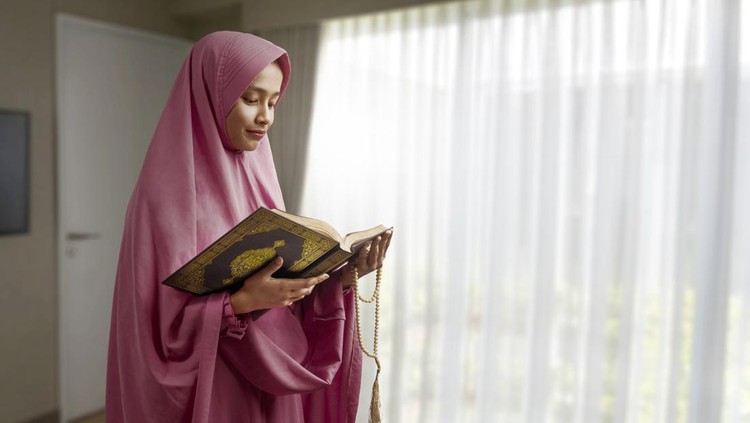 Asian Muslim woman in a veil holding prayer beads and reading the Quran at home