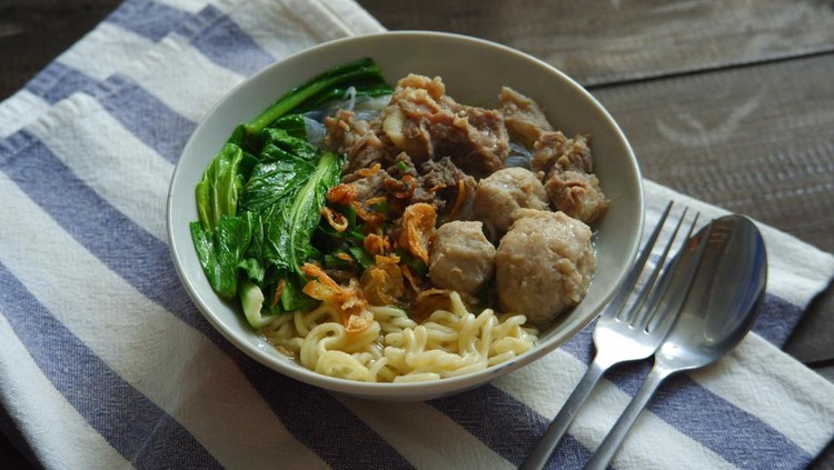 A bowl of noodles meatball soup and vegetables against wooden background