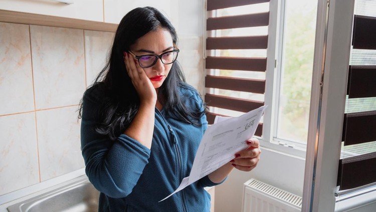 Portrait of a mid adult woman checking her energy bills at home, sitting in her bedroom. She has a worried expression and touches her face with her hand while looking at the bills. Focus on the woman while the interior architecture of the bedroom is defocused.