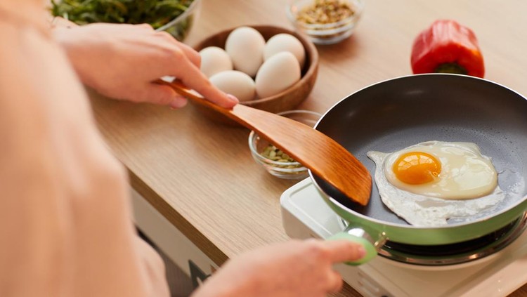 Warm-toned close up of unrecognizable young woman cooking eggs on frying pan while preparing healthy breakfast in morning, copy space