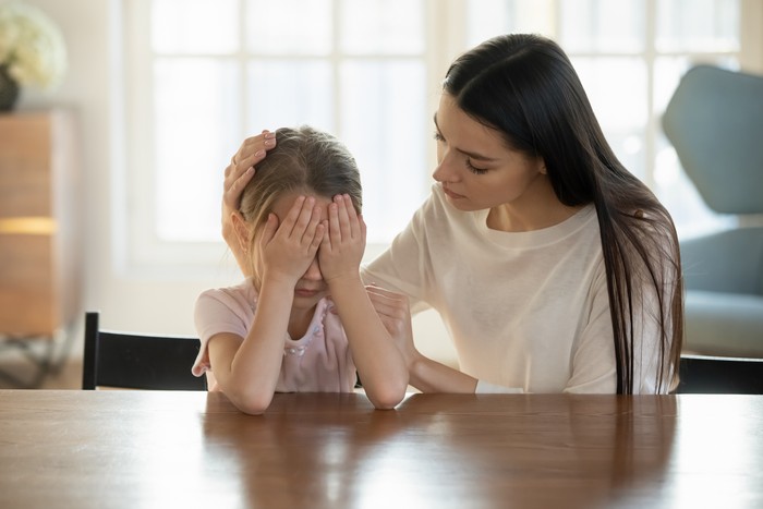 Caring mother calming and hugging crying upset little daughter, sitting at desk together, loving mum expressing support, comforting offended preschool girl, children psychologist concept