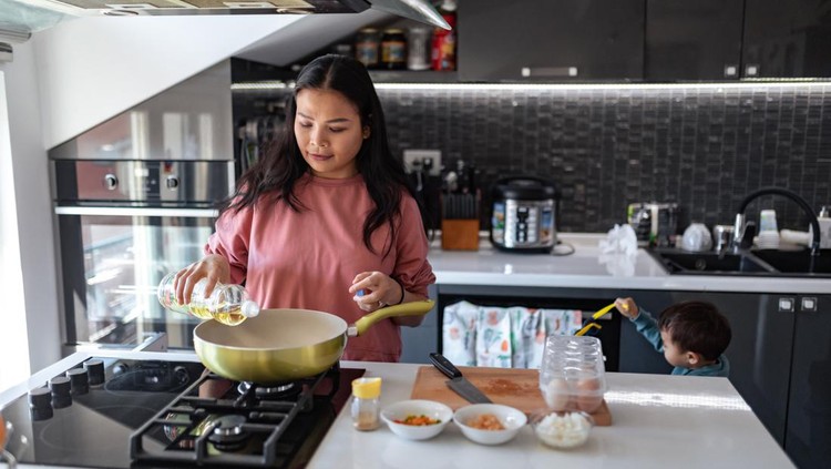 A hardworking mother prepares lunch. She pours olive oil into the pan while her little son plays in the background.