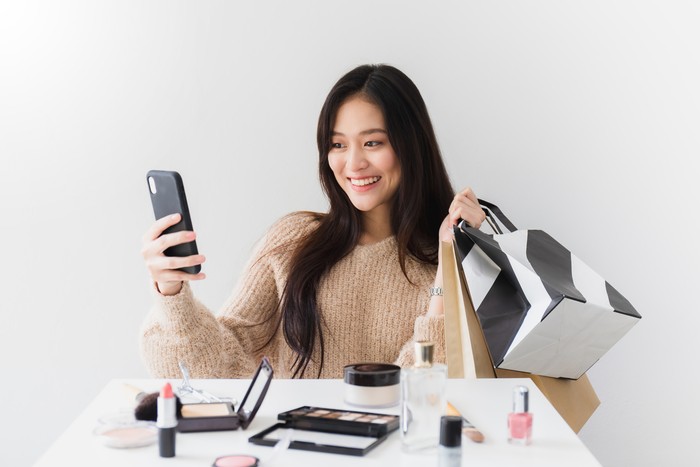 Asian beautiful women blogger are using the smartphone live streaming online with a shopping bag In the white room with cosmetics on table.Concept of online shopping business. Asian beautiful women blogger are using the smartphone live streaming online with a shopping bag In the white room with cosmetics on table.Concept of online shopping business.