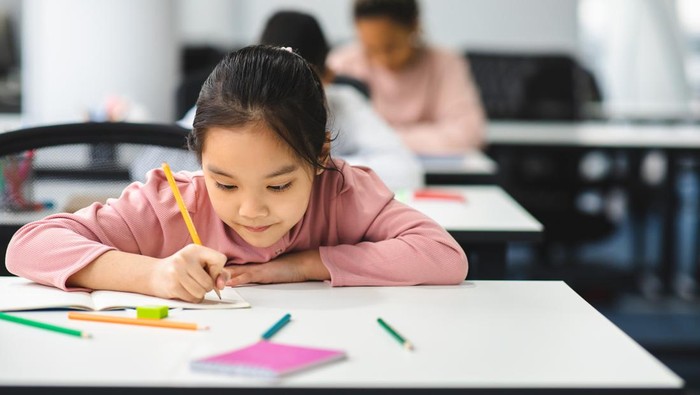 Tuition Concept. Portrait of concentrated small asian girl sitting at table in classroom, writing or drawing in notebook, doing homework or taking notes during lesson, selective focus, free copy space
