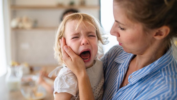 A mother holding a crying toddler daughter indoors in kitchen when cooking.