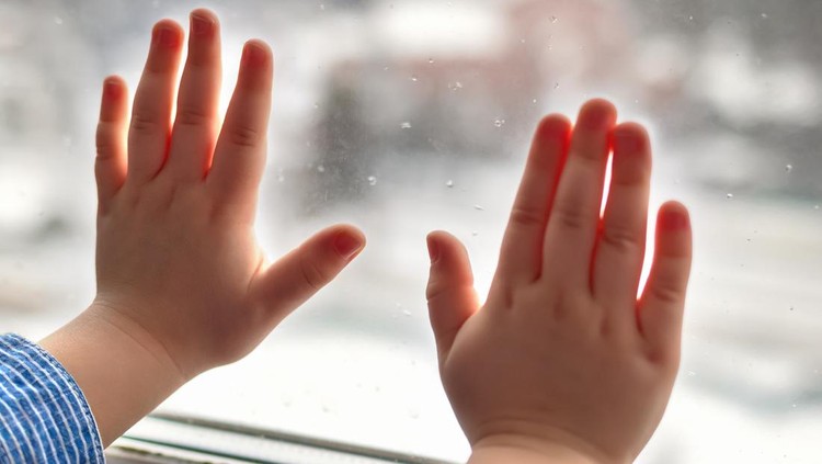 Close up and soft focus of hands of small child leaning against glass in window with snowflakes on winter day. Concept of child loneliness, sadness, waiting for parents.