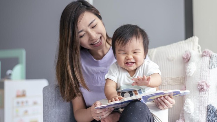 A beautiful young mother of Asian descent sits on the couch at home and affectionately holds her cute baby in her lap while reading a book to the child. The happy baby is looking with interest and delight at the book.