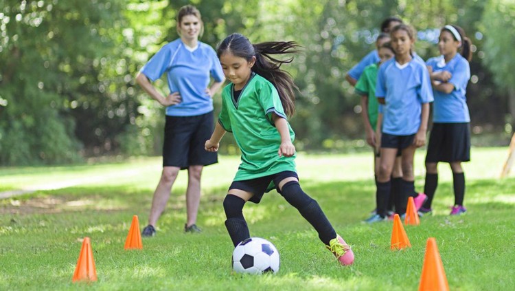 Asian elementary age soccer athlete confidently kicks the ball around orange practice cones. She is wearing a green uniform. Players in blue uniforms are lined up behind her as well as her young adult Caucasian female coach.