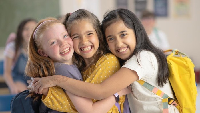 Three elementary school girls laugh and smile as the hug each other and look at the camera. They are wearing their backpacks and standing in a classroom.