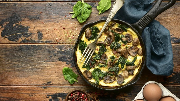 1138257405 Omelette with mushrooms and spinach in a cast iron pan on a dark rustic wooden background.Top view with copy space.