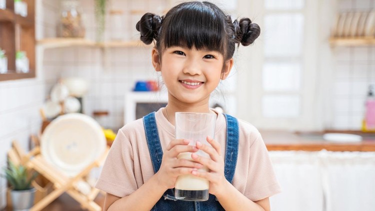 Portrait of Asian little kid holding a cup of milk in kitchen at home. Young preschool cute child girl or daughter stay home with smiling face, feel happy enjoy drinking milk and then look at camera.