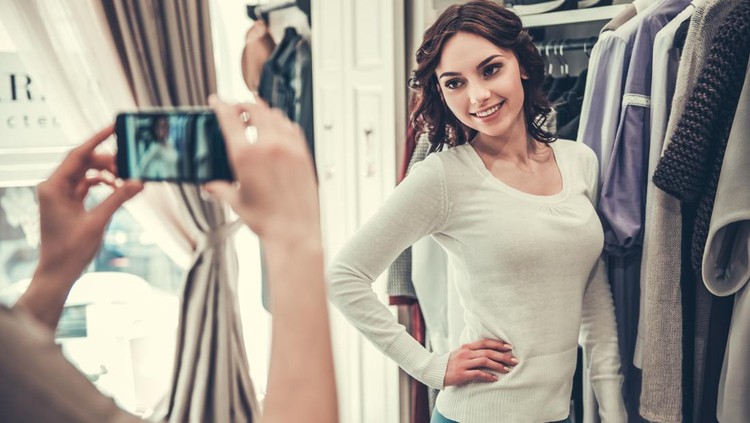 686762694 Girls in boutique. Attractive girl is trying on new clothes and smiling while her friend is taking photo