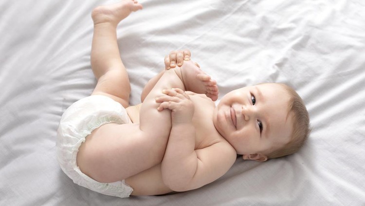 Ilustrasi Cara Memilih Popok Bayi yang Tepat Beautiful caucasian baby laying on bed, playing with feet and smiling at camera, top view, copy space. Adorable toddler in diaper having fun alone, lying on white, laughing. Toddler, baby, infant