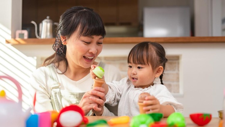 A Mother and her small daughter are playing together with toy vegetables and kitchen utensils in the living room at home.
