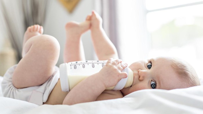 A Pretty baby girl drinks water from bottle lying on bed. Child weared diaper in nursery room.